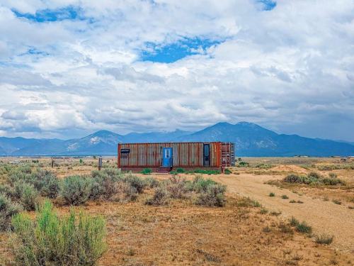 A modern container home in a desert landscape, surrounded by sparse vegetation and mountains under a cloudy sky.