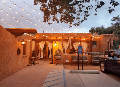 A cozy outdoor restaurant with string lights, adobe walls, and a patio area, set in the evening.