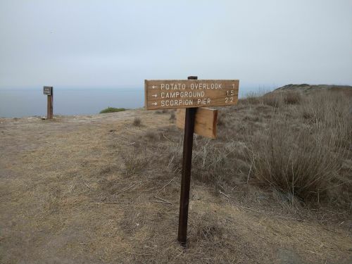 Wooden signpost with directions to Potato Overlook (15 miles) and Campground (22 miles) near a coastal area.
