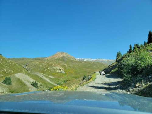 A gravel road winds through a mountainous landscape under a clear blue sky, with greenery and distant peaks visible.