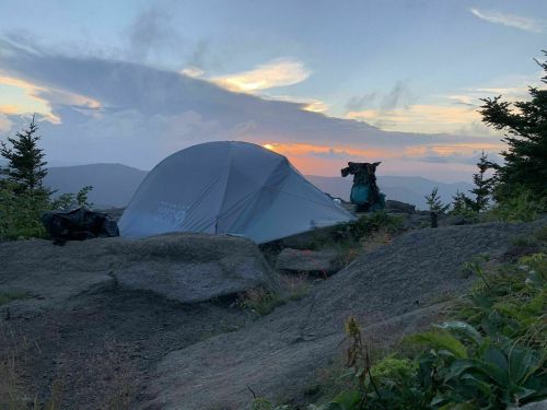 A tent set up on rocky terrain at sunset, surrounded by trees and mountains under a colorful sky.