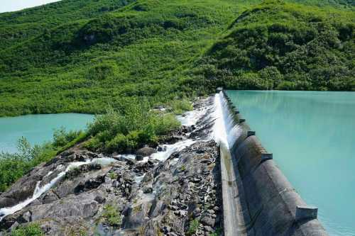 A dam with water cascading over rocks, surrounded by lush green hills and a turquoise lake.