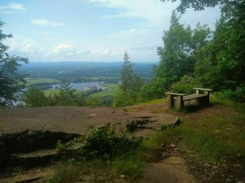 A scenic view from a rocky overlook, featuring a wooden bench surrounded by trees and rolling hills in the distance.