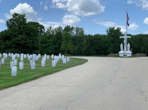 A serene cemetery with white gravestones lining a curved path, leading to a large cross and statues under a blue sky.