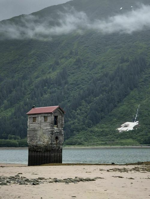 A weathered tower stands on stilts by a beach, surrounded by lush green mountains and a misty sky.