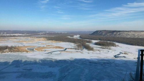 A snowy landscape with a winding river, surrounded by trees and hills under a clear blue sky.