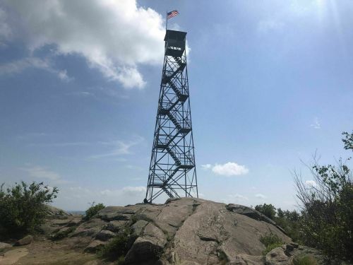 A tall metal observation tower on a rocky hill, with a flag flying at the top against a blue sky and scattered clouds.