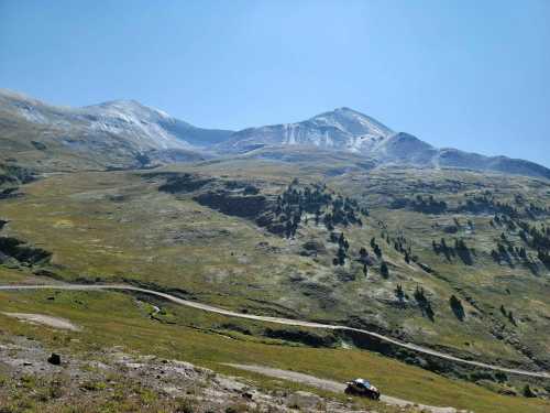 A scenic view of a mountainous landscape with green hills and a clear blue sky, featuring a winding dirt road and a small vehicle.