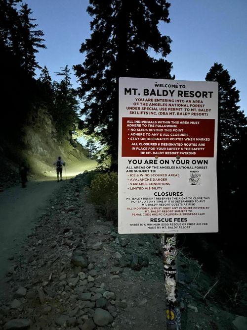 Sign at Mt. Baldy Resort warns visitors of safety rules and closures, with a hiker illuminated by a flashlight nearby.