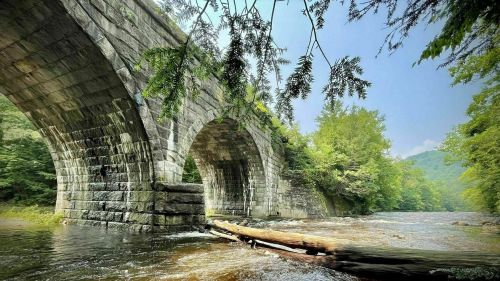A stone bridge arches over a river, surrounded by lush greenery and mountains in the background.