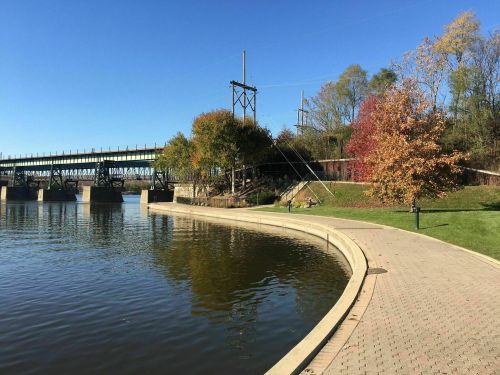 A serene riverside scene with a curved path, colorful trees, and a bridge in the background under a clear blue sky.