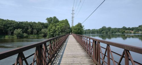 A wooden bridge stretches over a calm river, surrounded by lush greenery and power lines in the background.