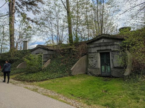 A person walks past two stone mausoleums covered in greenery, set in a peaceful outdoor environment.