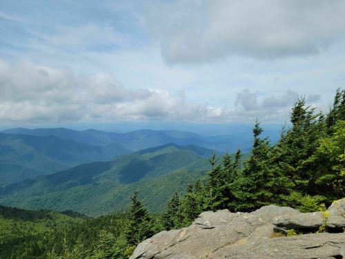 A panoramic view of lush green mountains under a cloudy sky, with rocky foreground and distant hills.