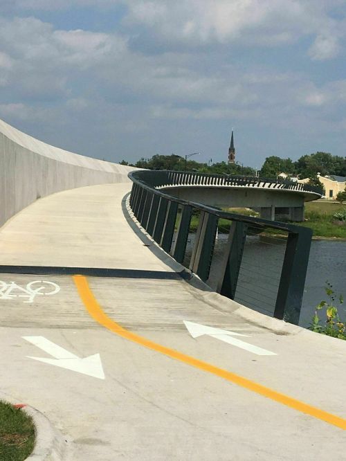 A winding bike path alongside a river, with a church steeple visible in the background under a partly cloudy sky.
