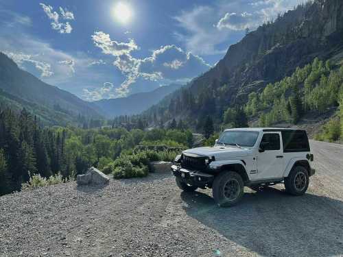 A white Jeep parked on a gravel road, surrounded by lush green trees and mountains under a bright blue sky.