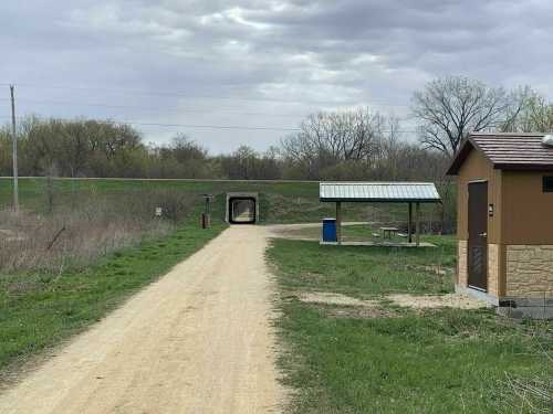 A dirt path leads to a tunnel, with a shelter and restroom nearby, surrounded by greenery under a cloudy sky.