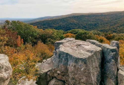 A rocky outcrop overlooks a colorful autumn landscape with rolling hills and trees in shades of orange and green.