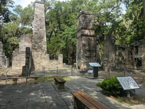 Ruins of stone structures surrounded by trees, with informational signs and benches in a historical site.
