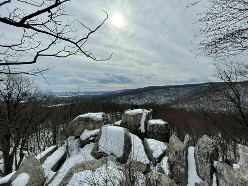 A snowy landscape with rocky outcrops under a cloudy sky, overlooking a distant valley and hills.
