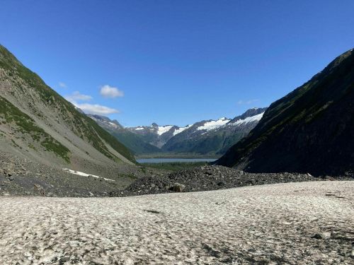 A scenic view of a valley surrounded by mountains, with a lake visible in the distance under a clear blue sky.