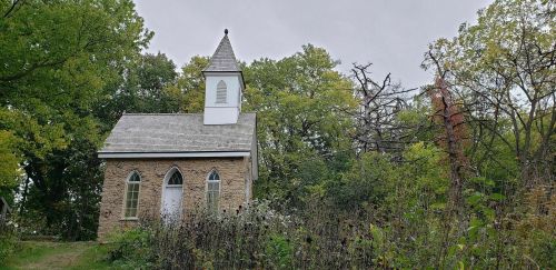 A small stone church with a steeple, surrounded by trees and overgrown vegetation under a cloudy sky.