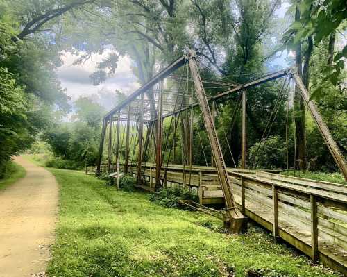 An old, rusty bridge stands beside a grassy path, surrounded by lush trees and a cloudy sky.