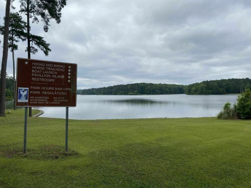 A serene lake view with a sign listing park regulations and amenities, surrounded by green grass and trees under a cloudy sky.