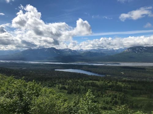 A panoramic view of mountains and a lake surrounded by lush greenery under a partly cloudy sky.