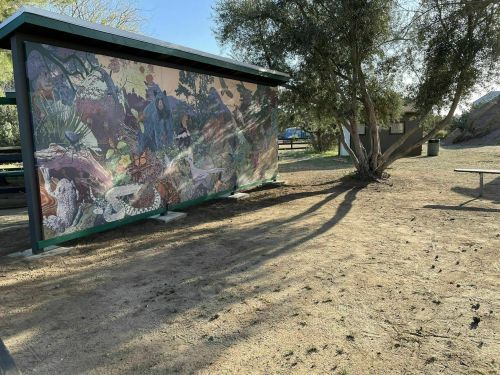 A colorful mural on a shelter in a park, surrounded by trees and a sandy area.