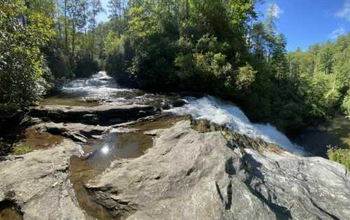 A serene waterfall cascades over rocky terrain, surrounded by lush green trees under a clear blue sky.