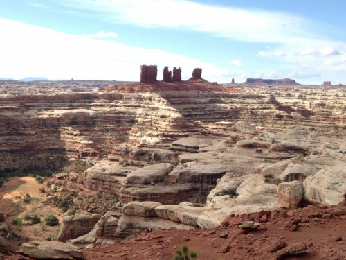 A vast desert landscape with layered rock formations and distant mesas under a blue sky with scattered clouds.