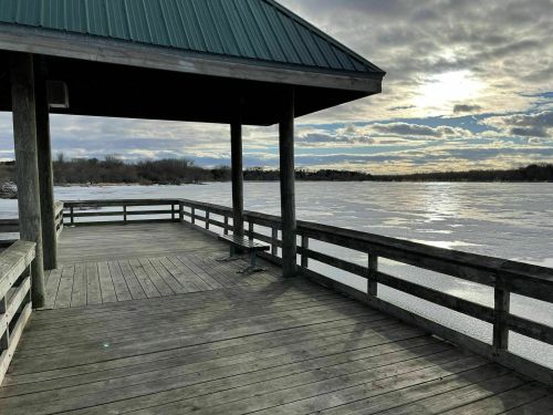 A wooden dock extends over a frozen lake under a cloudy sky, with sunlight peeking through the clouds.