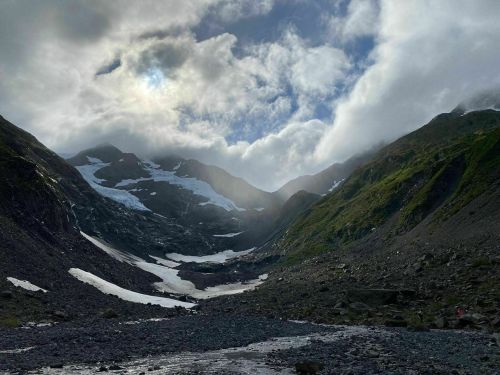 A rocky valley surrounded by mountains, with patches of snow and clouds partially covering the sun.