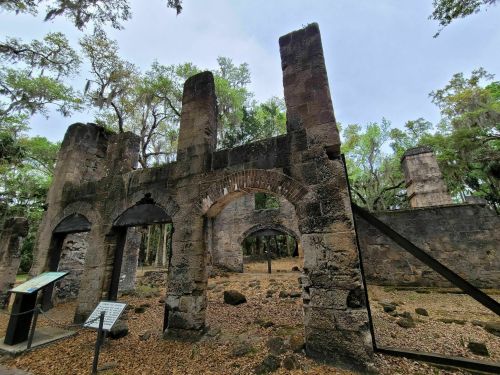 Ruins of a stone structure surrounded by trees and fallen leaves, with informational signs nearby.