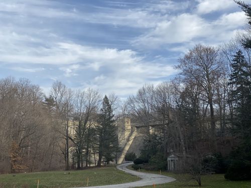 A winding road leads to a stone bridge surrounded by trees under a cloudy sky.