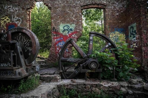 Abandoned industrial site with large gears and brick walls covered in graffiti, surrounded by overgrown vegetation.