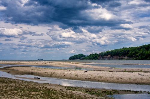 A serene beach scene with sandy shores, calm water, and dramatic clouds overhead, framed by green cliffs in the distance.