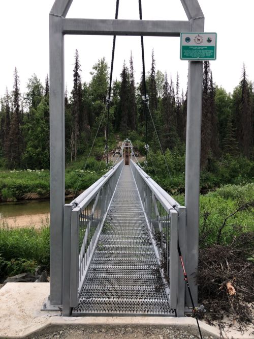 A metal suspension bridge spans a river, surrounded by trees and greenery, with a sign at the entrance.