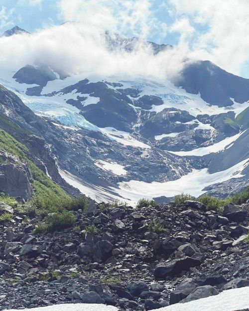 A stunning mountain landscape featuring glaciers, rocky terrain, and clouds hovering over the peaks.