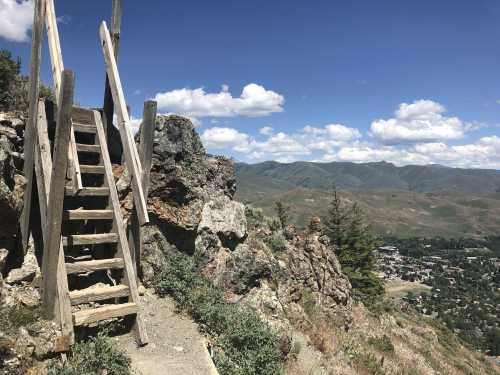 A wooden staircase leads up a rocky hillside, overlooking a valley and distant mountains under a blue sky with clouds.