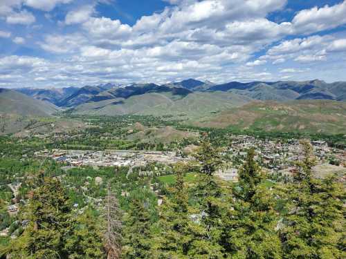 A panoramic view of a valley surrounded by mountains, with lush greenery and a small town visible below under a cloudy sky.