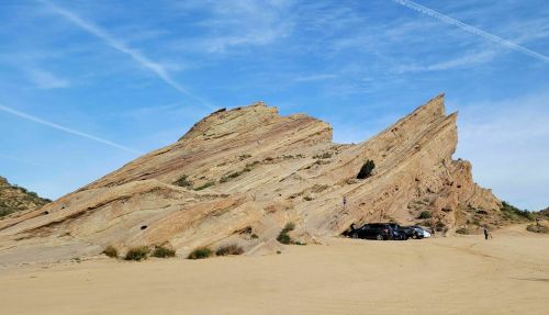 A striking rock formation rises above a sandy area, with a few cars parked nearby under a clear blue sky.