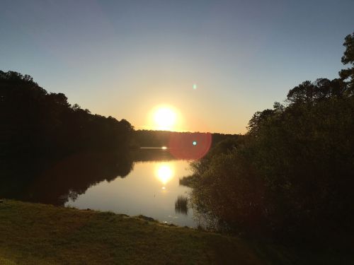 A serene sunset over a calm lake, with trees lining the shore and a clear sky. Reflections shimmer on the water.