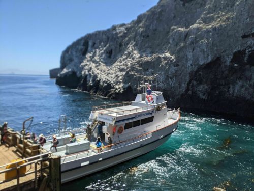 A boat docked at a rocky shore with clear blue water and people boarding on a sunny day.