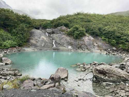 A serene turquoise pond surrounded by rocky terrain and lush greenery, with a small waterfall in the background.