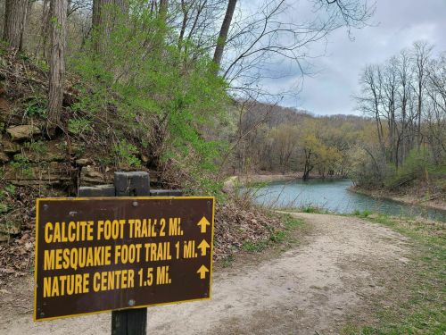 Signpost indicating trail distances: Calcite Foot Trail 2 mi, Mesquakie Foot Trail 1 mi, Nature Center 1.5 mi, near a river.