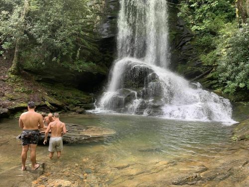 Three people stand near a waterfall, surrounded by lush greenery and a clear pool of water.