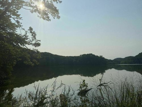 A serene lake surrounded by lush greenery, reflecting the sun and hills under a clear sky.