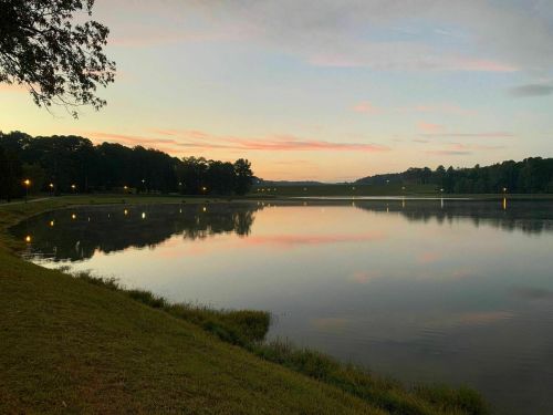 A serene lake at sunset, reflecting soft colors in the water, surrounded by trees and lights along the shore.